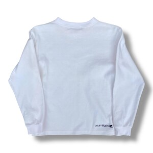 May include: A white long-sleeve shirt with a crew neck. The shirt has a minimalist design and features the brand name "courr&egrave;ges" in black text on the lower right sleeve. The shirt is made of a soft, comfortable material.