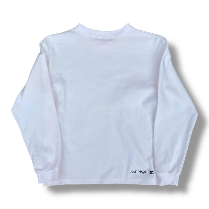 May include: A white long-sleeve shirt with a crew neck. The shirt has a minimalist design and features the brand name "courr&egrave;ges" in black text on the lower right sleeve. The shirt is made of a soft, comfortable material.