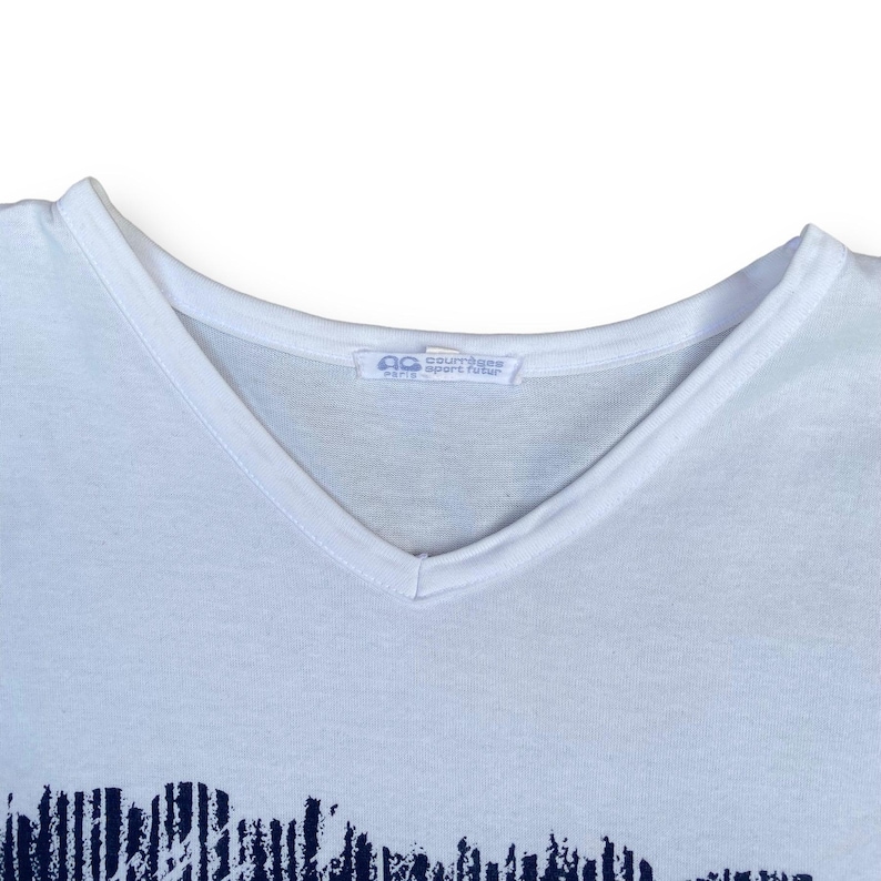 K&ouml;nnte beinhalten: Wei&szlig;es T-Shirt mit V-Ausschnitt und einem schwarz-wei&szlig;en Grafikdruck. Das Shirt hat ein Etikett mit der Aufschrift "Courr&egrave;ges Sport Rider Paris".