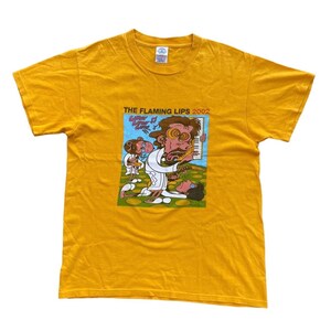 Puede incluir: Camiseta amarilla con una ilustración de dibujos animados de una banda, The Flaming Lips, tocando música. El texto "THE FLAMING LIPS 2002" está impreso en la camiseta.