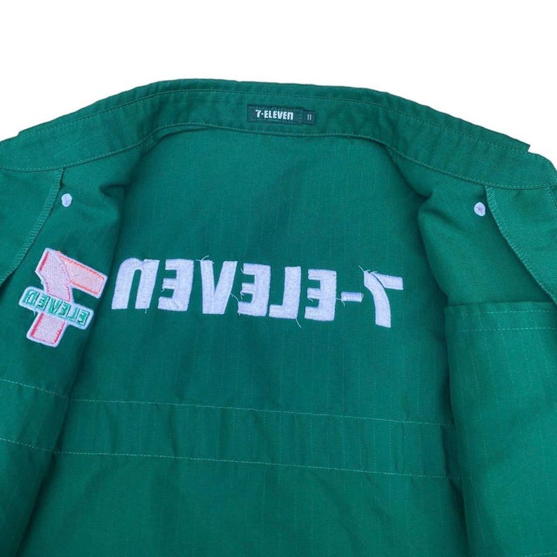 May include: Green work shirt with white text and a 7-Eleven logo patch. The shirt features the 7-Eleven logo on the left chest and the 7-Eleven text across the back. The shirt has a button-down front and a collar.
