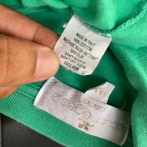 Puede incluir: Una etiqueta blanca con texto negro en una camiseta verde. La etiqueta dice "Made in Italy, 100% algod&oacute;n, Lavado a m&aacute;quina 40&deg;C/104&deg;, Secar en plano, No usar lej&iacute;a, No limpiar en seco, Planchar en fr&iacute;o".