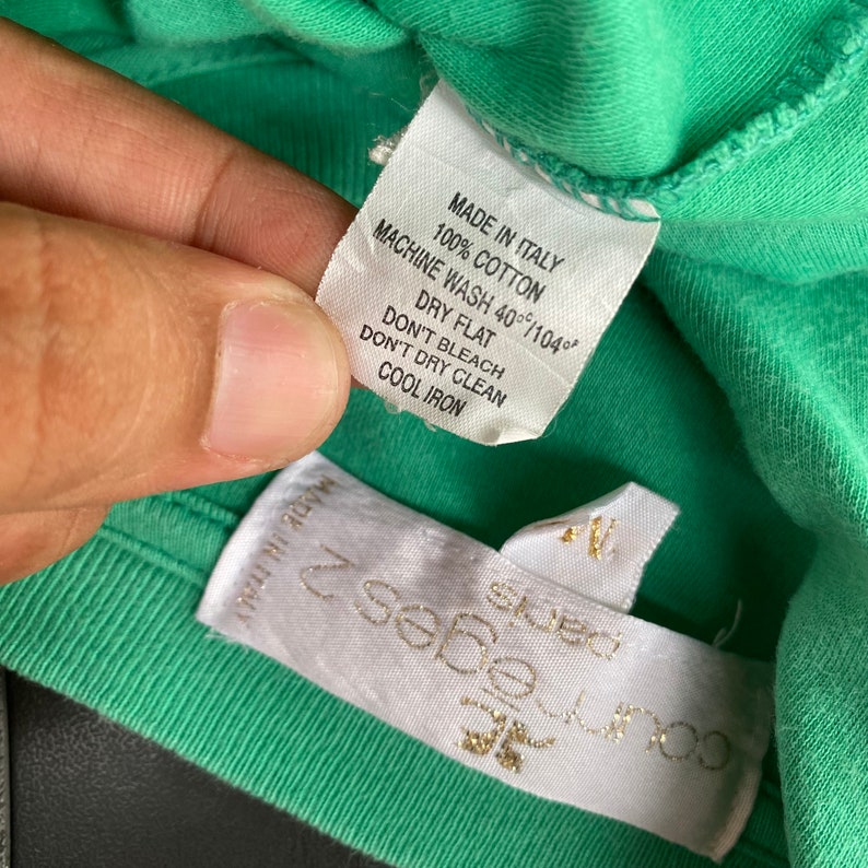Puede incluir: Una etiqueta blanca con texto negro en una camiseta verde. La etiqueta dice "Made in Italy, 100% algod&oacute;n, Lavado a m&aacute;quina 40&deg;C/104&deg;, Secar en plano, No usar lej&iacute;a, No limpiar en seco, Planchar en fr&iacute;o".