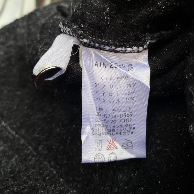 K&ouml;nnte beinhalten: Nahaufnahme eines wei&szlig;en Etiketts mit schwarzem Text und einem silbernen Metallring. Das Etikett enth&auml;lt den Text "ATN-2059 W 36" und japanischen Text, der die Materialzusammensetzung angibt: Acryl 70 %, Nylon 15 % und Polyester 15 %.