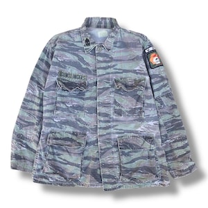 Puede incluir: Chaqueta de camuflaje vintage en tonos verdes, grises y azules. La chaqueta tiene cuatro bolsillos delanteros, cuello y un parche con una calavera y las letras "VR". La palabra "GUNSLINGERS" está impresa encima del bolsillo izquierdo del pecho.