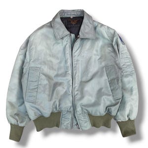 May include: A pale blue bomber jacket with a collared neckline, two front pockets, and olive green ribbed cuffs and hem. The jacket has a zip closure and a patch on the upper arm. A vintage style jacket.