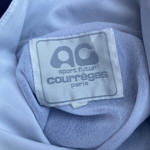 May include: Close-up of a light blue garment tag with the text "ac sport futur courr&egrave;ges paris". The tag is rectangular with rounded corners and a light grey border. The background is a light blue fabric.