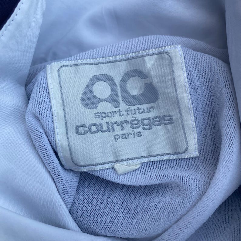May include: Close-up of a light blue garment tag with the text "ac sport futur courr&egrave;ges paris". The tag is rectangular with rounded corners and a light grey border. The background is a light blue fabric.