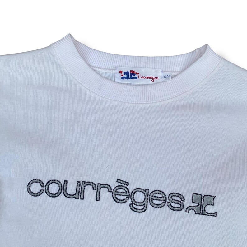 May include: A white crew neck sweatshirt with the brand name "courr&egrave;ges" in a dark gray, stylized font across the chest. The brand's logo is visible on the collar. The sweatshirt is made of a soft material.
