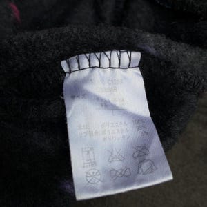 Puede incluir: Primer plano de una etiqueta de ropa blanca con texto negro y s&iacute;mbolos de cuidado. La etiqueta est&aacute; adherida a una tela gris oscuro con toques de rosa. Contiene informaci&oacute;n sobre los materiales y la talla de la prenda.