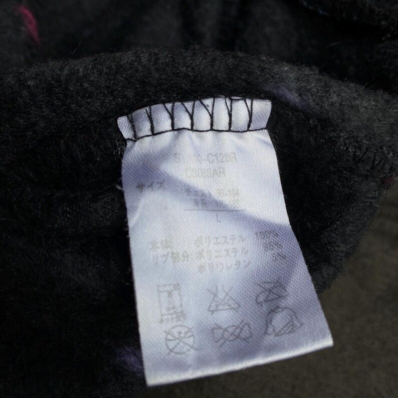 Puede incluir: Primer plano de una etiqueta de ropa blanca con texto negro y s&iacute;mbolos de cuidado. La etiqueta est&aacute; adherida a una tela gris oscuro con toques de rosa. Contiene informaci&oacute;n sobre los materiales y la talla de la prenda.