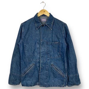 Könnte beinhalten: Eine blaue Jeans-Arbeitsjacke mit Knopfleiste, zwei Brusttaschen und zwei unteren Taschen. Die Jacke hat ein Etikett mit der Aufschrift "Blue Blue" auf der linken Brusttasche.
