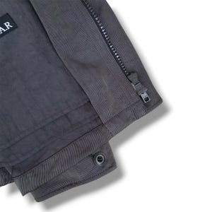 K&ouml;nnte beinhalten: Eine dunkelgraue Jacke mit vertikalem Rei&szlig;verschluss und Metallschieber, einem rechteckigen schwarzen Aufn&auml;her mit dem wei&szlig;en Text A.R oben links sowie einem einzelnen Knopfverschluss am unteren Saum des Kleidungsst&uuml;cks.