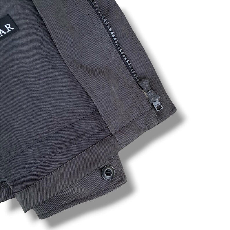 K&ouml;nnte beinhalten: Eine dunkelgraue Jacke mit vertikalem Rei&szlig;verschluss und Metallschieber, einem rechteckigen schwarzen Aufn&auml;her mit dem wei&szlig;en Text A.R oben links sowie einem einzelnen Knopfverschluss am unteren Saum des Kleidungsst&uuml;cks.