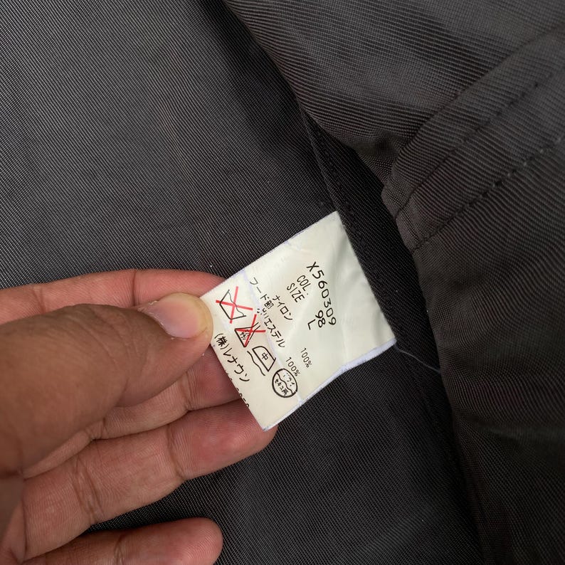 K&ouml;nnte beinhalten: Ein wei&szlig;es Pflegeetikett, das in ein dunkelgraues Stofffutter eingen&auml;ht ist. Das Etikett enth&auml;lt den Text X560309, COL 98, SIZE L und listet eine Zusammensetzung aus 100% Nylon und 100% Polyester sowie japanische Pflegesymbole und Anweisungen auf.