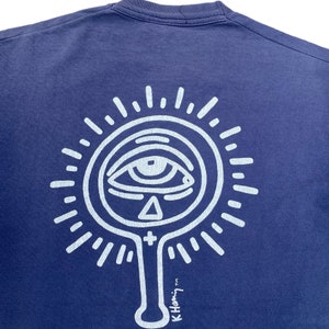 Puede incluir: Una camiseta azul marino con un gr&aacute;fico blanco de un ojo dentro de un c&iacute;rculo con rayos que emanan de &eacute;l. El gr&aacute;fico tambi&eacute;n incluye un tri&aacute;ngulo y una cruz. El texto "K.Hammy TM" est&aacute; en la parte inferior del gr&aacute;fico.