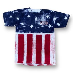 Pode incluir: Uma camiseta branca com uma estampa de bandeira americana desbotada. A camiseta tem um esquema de cores vermelho, branco e azul e apresenta o texto "Combat Force" em um círculo no peito.