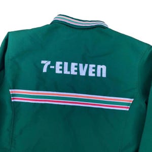 May include: A green jacket with the text "7-ELEVEN" in white block letters across the back. The jacket has a striped design in red, white, and orange near the bottom. The collar has a striped design in white, green, and red.