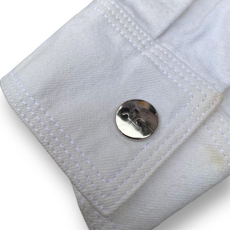 May include: Close-up of a white denim jacket cuff with a silver button featuring the letters "RC" in a logo.