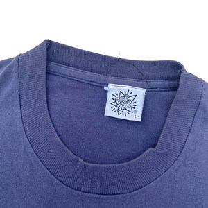 Puede incluir: Primer plano de una camiseta azul con una etiqueta blanca que dice "Pop Shop" en un dise&ntilde;o en forma de estrella. La etiqueta tambi&eacute;n tiene un peque&ntilde;o s&iacute;mbolo de marca registrada.