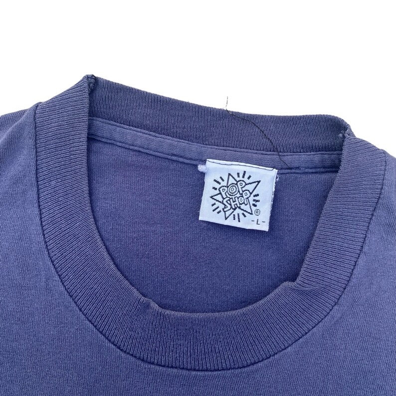 Puede incluir: Primer plano de una camiseta azul con una etiqueta blanca que dice "Pop Shop" en un dise&ntilde;o en forma de estrella. La etiqueta tambi&eacute;n tiene un peque&ntilde;o s&iacute;mbolo de marca registrada.