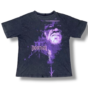 May include: Dark gray t-shirt featuring a purple graphic of a wrestler's face and the word "UNDERTAKER" in purple. The graphic has a distressed, faded look. The shirt has a crew neck and short sleeves.