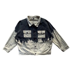 Könnte beinhalten: Jeansjacke mit einzigartigem Design. Die Jacke hat eine schwarze und helle Waschung mit einem dunklen, farbähnlichen Effekt auf der Oberseite. Sie hat zwei Brusttaschen und eine Knopfleiste.