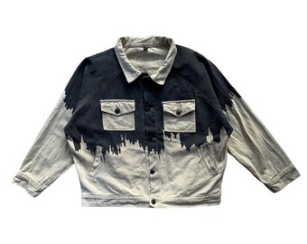 May include: A denim jacket with a unique design. The jacket features a black and light-colored wash, with a dark paint-like effect across the top. It has two chest pockets and a button-up front.