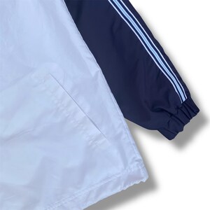 May include: A white and navy blue jacket with a pocket and a navy blue sleeve. The sleeve has two light blue stripes. The jacket is made of a lightweight material.