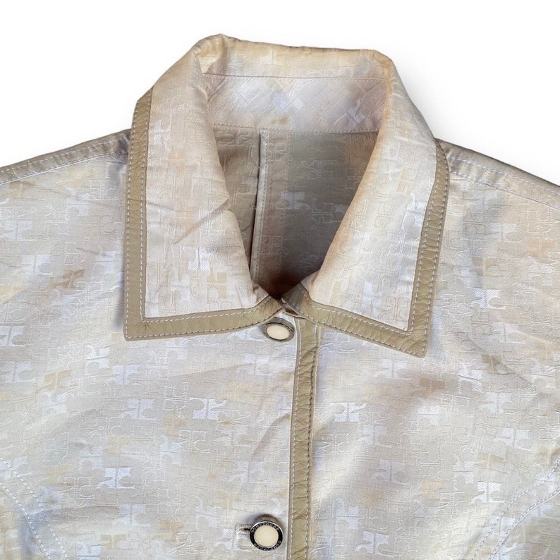 May include: A beige jacket with a white patterned design and a brown leather collar. The jacket has two buttons on the front.