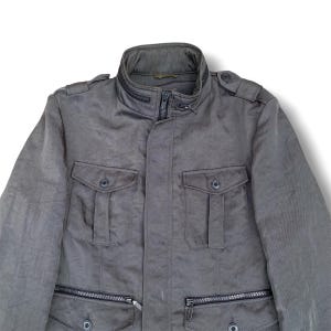 K&ouml;nnte beinhalten: Eine dunkelgraue Utility-Jacke mit hohem Kragen und Rei&szlig;verschluss-Detail, zwei gekn&ouml;pften Brusttaschen und zwei unteren Rei&szlig;verschlusstaschen. Die Jacke verf&uuml;gt &uuml;ber Schulterklappen und eine mittige Frontleiste &uuml;ber einem Rei&szlig;verschluss.
