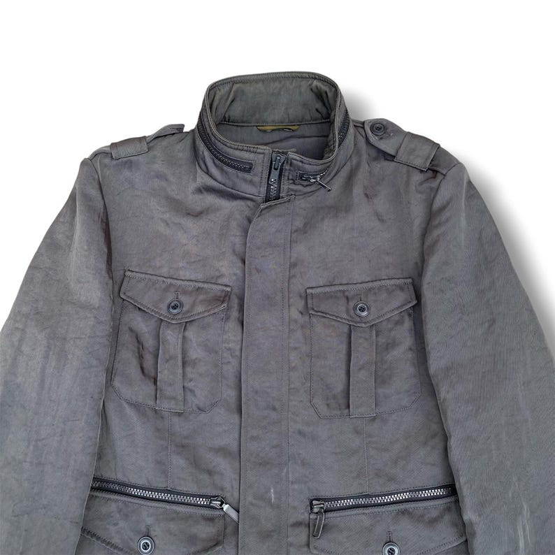 K&ouml;nnte beinhalten: Eine dunkelgraue Utility-Jacke mit hohem Kragen und Rei&szlig;verschluss-Detail, zwei gekn&ouml;pften Brusttaschen und zwei unteren Rei&szlig;verschlusstaschen. Die Jacke verf&uuml;gt &uuml;ber Schulterklappen und eine mittige Frontleiste &uuml;ber einem Rei&szlig;verschluss.