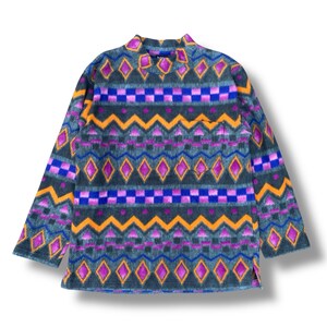 May include: A long-sleeved fleece shirt with a vibrant geometric pattern. The design features repeating diamond, zigzag, and square shapes in shades of purple, orange, and blue against a dark teal background. The shirt has a small chest pocket.