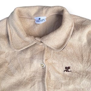 May include: A beige fleece button-up shirt with a brown embroidered logo that says "Courr&egrave;ges" on the collar and a brown embroidered logo on the front that says "JC".