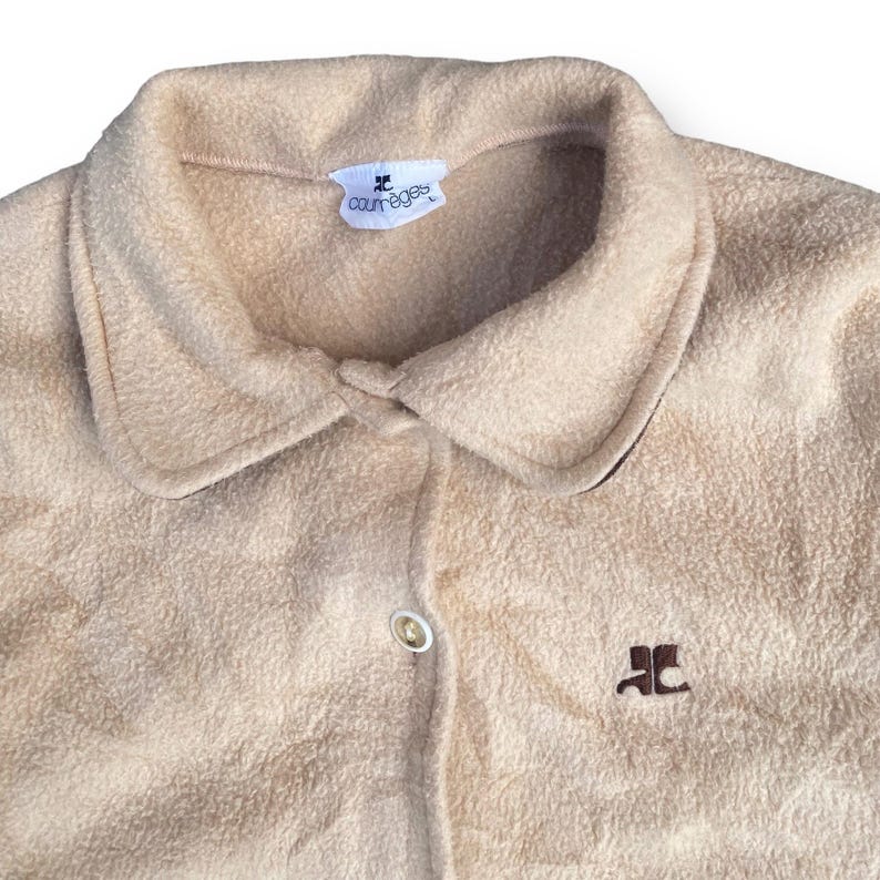 May include: A beige fleece button-up shirt with a brown embroidered logo that says "Courr&egrave;ges" on the collar and a brown embroidered logo on the front that says "JC".