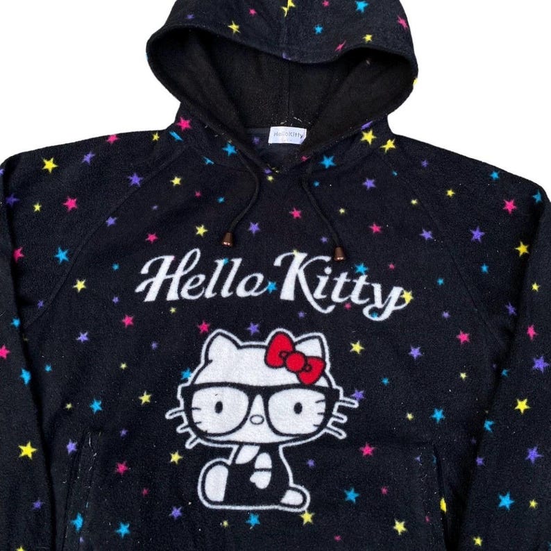Puede incluir: Sudadera con capucha negra con un estampado de Hello Kitty y un patr&oacute;n de estrellas de colores. El estampado de Hello Kitty es blanco con un lazo rojo y gafas. El texto "Hello Kitty" est&aacute; en escritura blanca.