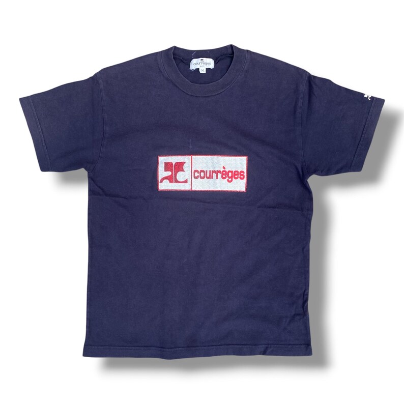 K&ouml;nnte beinhalten: Ein dunkelblaues Kurzarm-T-Shirt mit einer rechteckigen Grafik auf der Brust auf wei&szlig;em Grund, einem roten Logo und dem Schriftzug "courr&egrave;ges" in Rot. Das T-Shirt hat einen Rundhalsausschnitt und ein kleines wei&szlig;es Logo am linken &Auml;rmel.