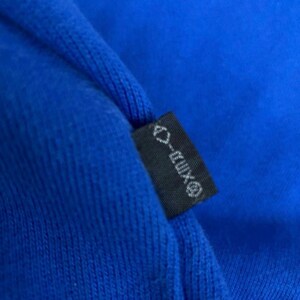 Peut inclure: Gros plan sur un tissu bleu roi avec une petite &eacute;tiquette noire. L'&eacute;tiquette porte un texte blanc et un logo. Le tissu semble &ecirc;tre un mat&eacute;riau tricot&eacute;.