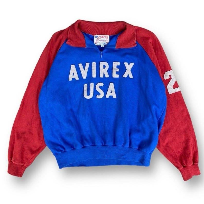 Peut inclure: Un sweat-shirt vintage bleu, rouge et blanc Avirex USA. Le sweat-shirt a un devant bleu avec les mots "AVIREX USA" en lettres blanches. Les manches sont rouges et le chiffre "2" est sur la manche droite.