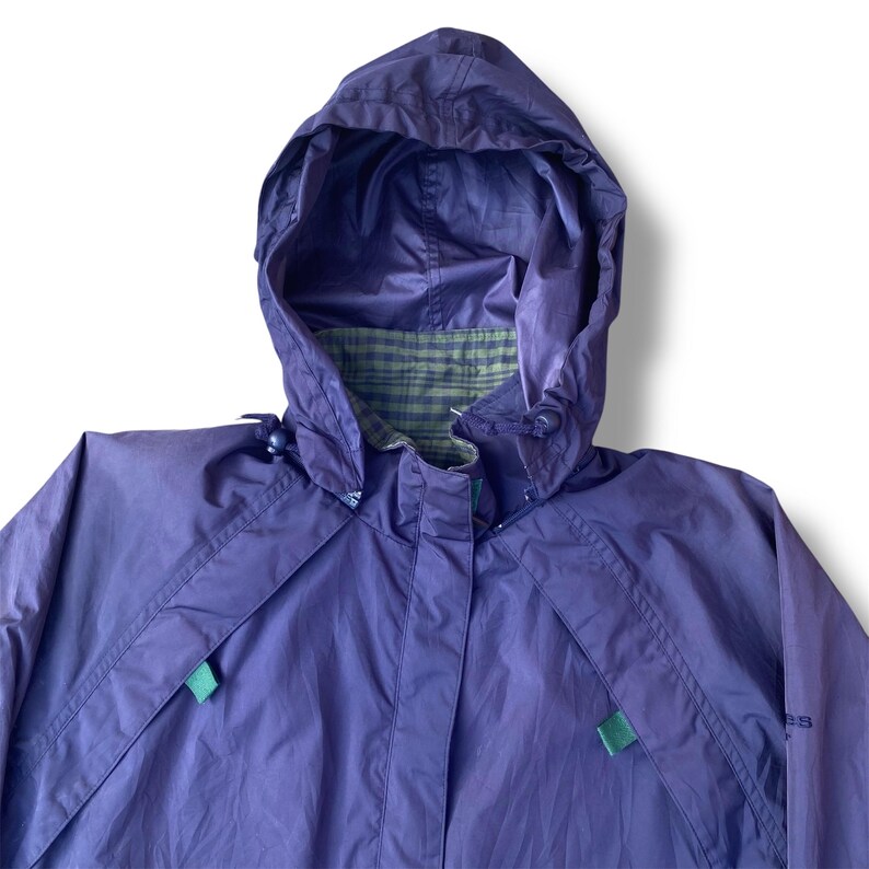 May include: A purple hooded rain jacket with a green plaid lining visible at the hood's opening. The jacket has a zipper closure and green accents on the front. The jacket is made of a lightweight, water-resistant material.