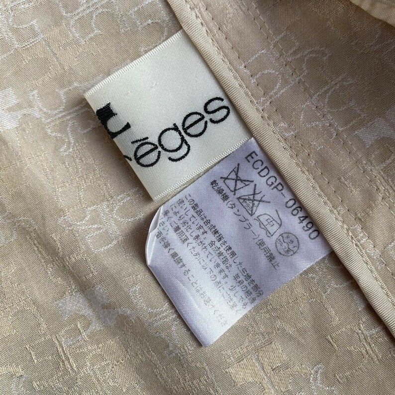 May include: A white tag with the word "&egrave;ges" printed in black. The tag is attached to a beige fabric with a white pattern. The tag also includes care instructions in Japanese.