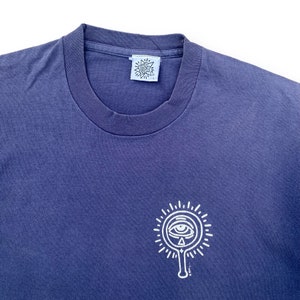 Puede incluir: Una camiseta azul oscuro con un gr&aacute;fico blanco de un ojo estilizado dentro de un c&iacute;rculo con rayos que emanan de &eacute;l. La camiseta tiene una etiqueta blanca con el texto "The Great Divide" en ella.