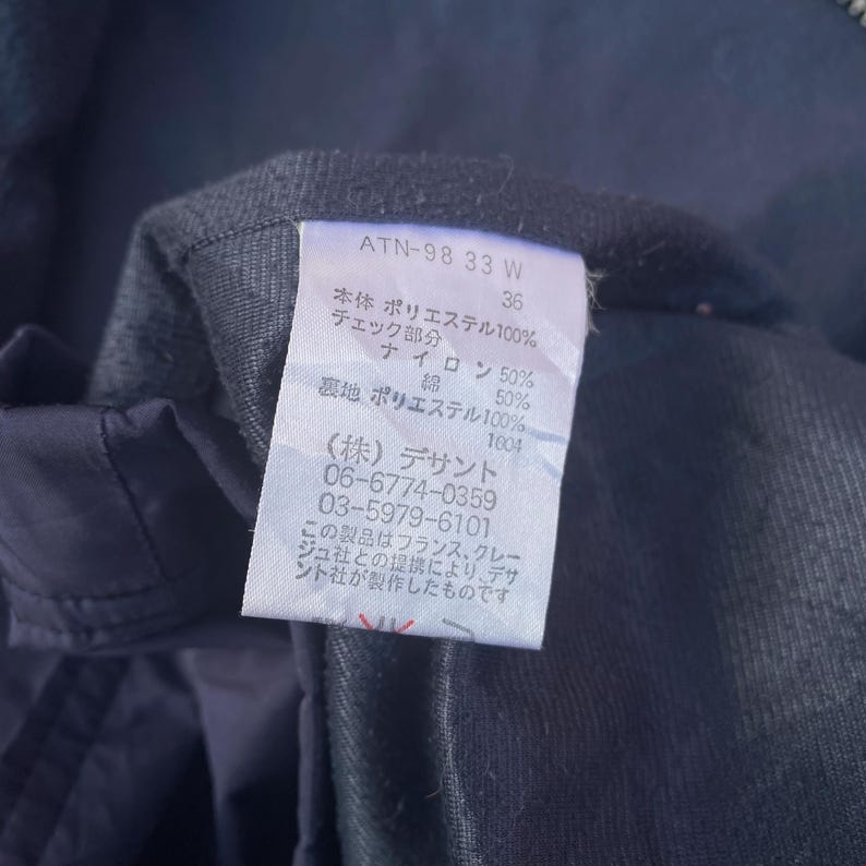 May include: Close-up of a garment tag with Japanese text and the code ATN-98 33 W. The tag also lists fabric composition: 100% polyester, 50% nylon, and 50% cotton. The tag is white with black text.