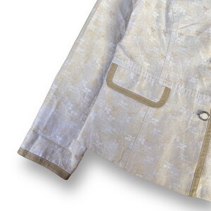 May include: A beige blazer with a white patterned design and brown trim. The blazer has a single button closure and a pocket on the left side.