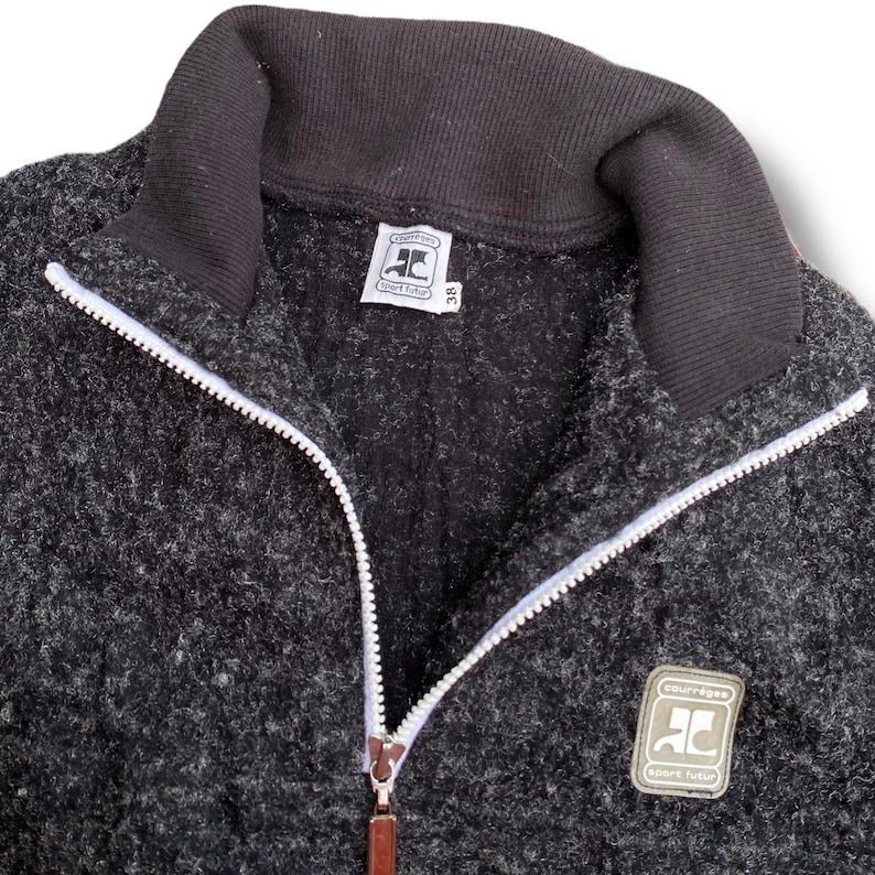 K&ouml;nnte beinhalten: Dunkelgraue Jacke mit Rei&szlig;verschluss, schwarzem Kragen und wei&szlig;em Rei&szlig;verschluss. Die Jacke hat einen kleinen Logoaufn&auml;her und ein Etikett mit dem Text "courr&egrave;ges sport futur".