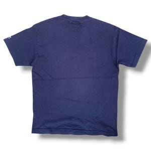 K&ouml;nnte beinhalten: Ein einfarbiges, marineblaues T-Shirt mit kurzen &Auml;rmeln und Rundhalsausschnitt. Das Kleidungsst&uuml;ck weist ein kleines wei&szlig;es Logodetail am Rand des linken &Auml;rmels auf. Der Stoff wirkt wie ein Standard-Baumwollstrick mit schlichtem Design.