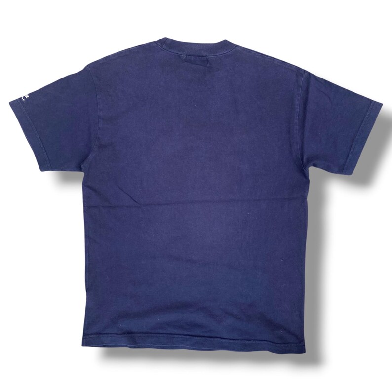 K&ouml;nnte beinhalten: Ein einfarbiges, marineblaues T-Shirt mit kurzen &Auml;rmeln und Rundhalsausschnitt. Das Kleidungsst&uuml;ck weist ein kleines wei&szlig;es Logodetail am Rand des linken &Auml;rmels auf. Der Stoff wirkt wie ein Standard-Baumwollstrick mit schlichtem Design.