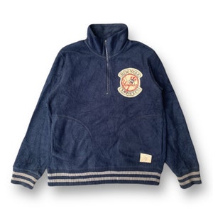 Könnte beinhalten: Marineblaues Fleece-Pullover mit hohem Kragen und einem Viertel-Reißverschluss. Mit dem New York Yankees Logo-Patch auf der Brust. Der Saum und die Ärmelbündchen haben grau-weiße Streifen. Ein kleines Etikett ist unten angenäht.