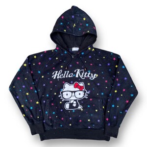 Puede incluir: Sudadera con capucha negra con un gr&aacute;fico de Hello Kitty con gafas y un lazo rojo. La sudadera est&aacute; cubierta de patrones de estrellas de colores y las palabras "Hello Kitty" est&aacute;n impresas encima del gr&aacute;fico.