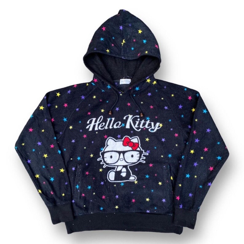 Puede incluir: Sudadera con capucha negra con un gr&aacute;fico de Hello Kitty con gafas y un lazo rojo. La sudadera est&aacute; cubierta de patrones de estrellas de colores y las palabras "Hello Kitty" est&aacute;n impresas encima del gr&aacute;fico.